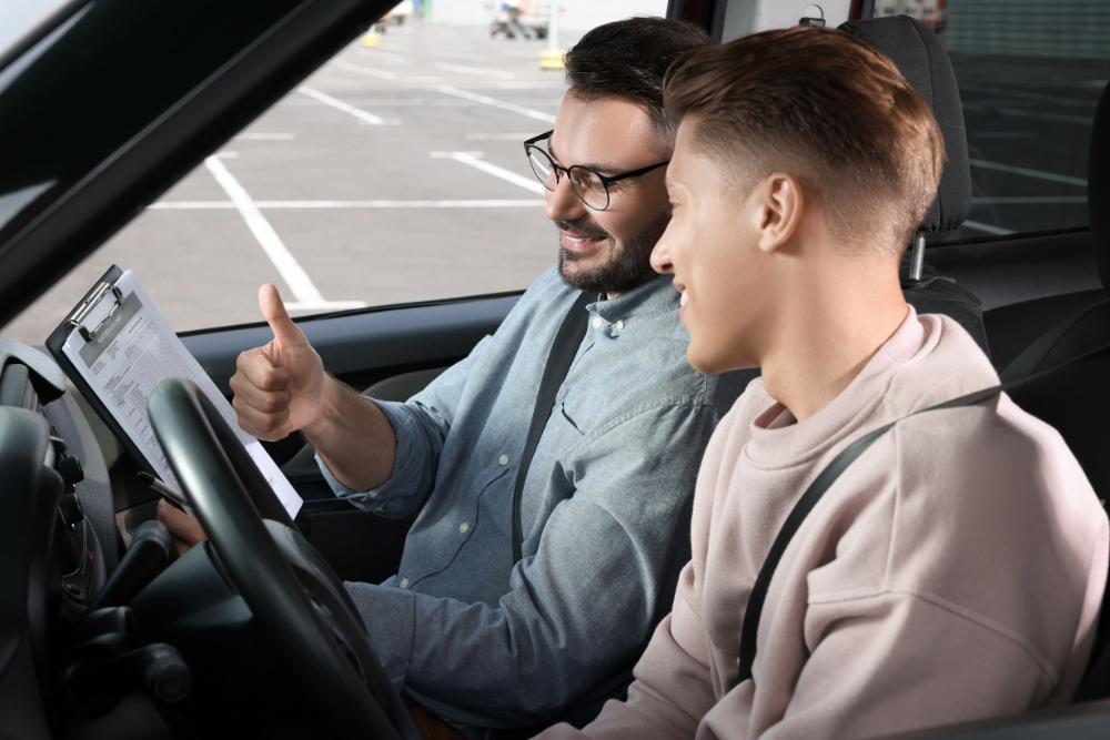 Un moniteur d’auto-école souriant montre un pouce levé à un jeune élève conducteur, à bord d’une voiture stationnée sur un parking, avec une feuille d’évaluation sur un clipboard.