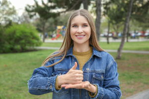 Jeune femme souriante utilisant la langue des signes française (LSF) dans un parc, réalisant un geste signifiant "bien" ou "ok" avec les mains.