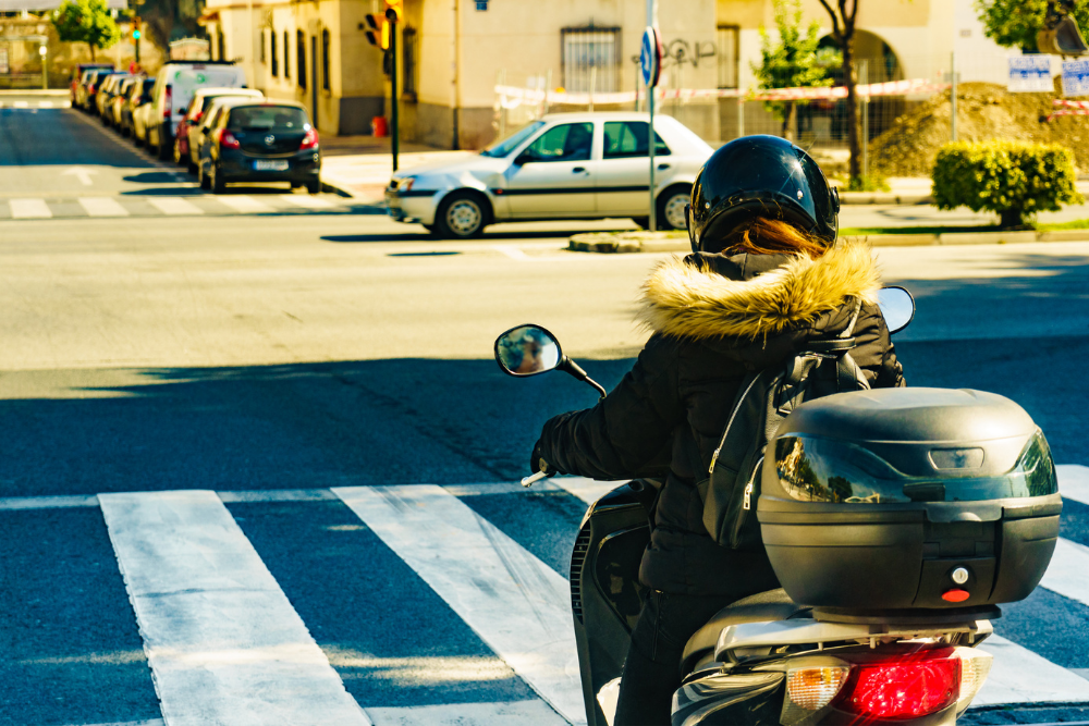 Motard(e) vu(e) de dos, à l’arrêt à un passage piéton en milieu urbain, portant un casque et un blouson avec capuche, prêt(e) à redémarrer.