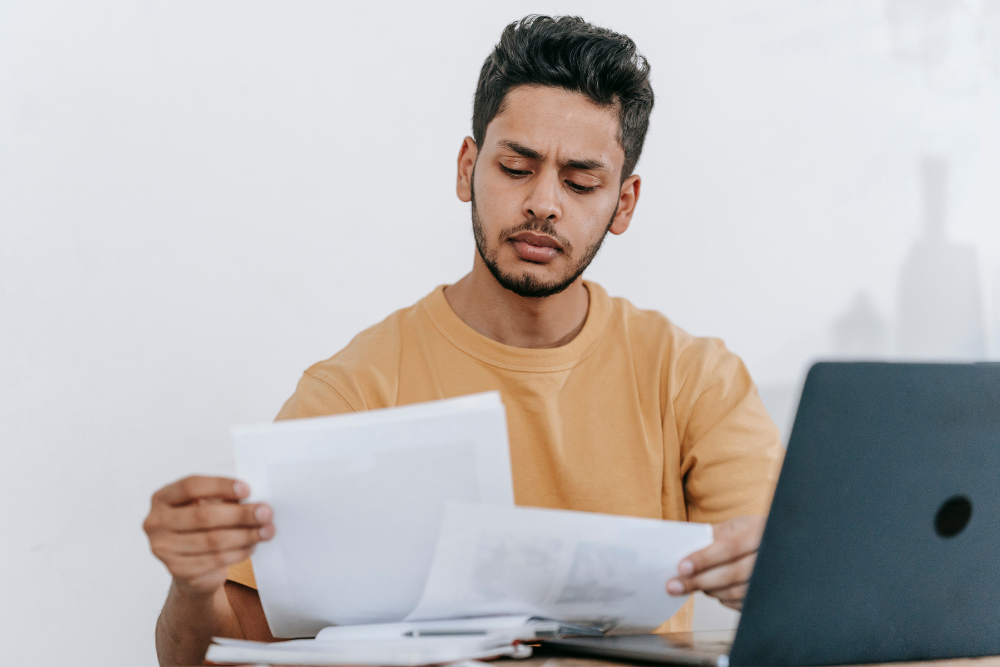 Un jeune homme examine des documents administratifs pour changer d'auto-école, assis à un bureau avec un ordinateur portable ouvert devant lui.